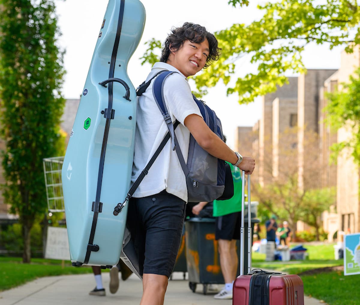 A smiling male student carries a light blue cello case on his back, a backpack, and pulls a red suitcase, walking on campus during move-in.